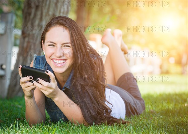 Attractive happy mixed-race young female laying in the grass texting on her cell phone outside
