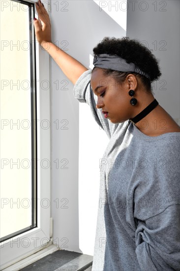 Eine nachdenkliche Afro-Amerikanische junge huebsche Frau steht an einem Fenster und blickt nachdenklich zu Boden. Sie traegt einen grauen Pullover mit einem passenden Stirnband
