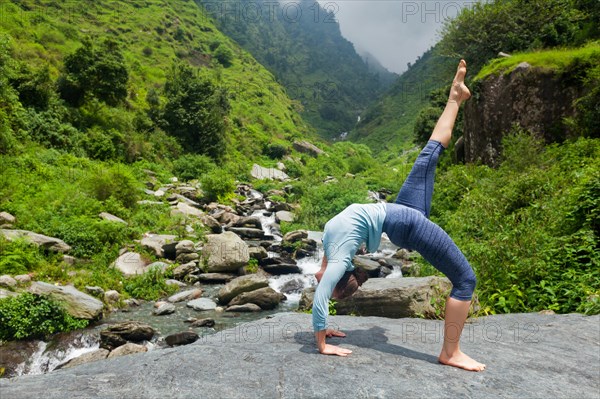 Woman doing yoga asana eka pada urdva dhanurasana Upward Bow Pose outdoors at waterfall in Himalayas