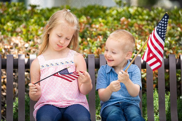 Young sister and brother comparing each others american flag size on the bench at the park