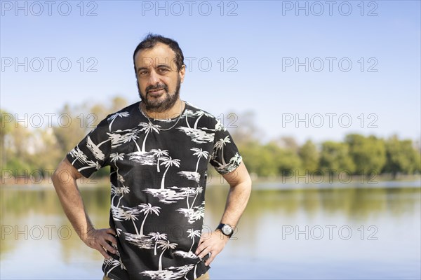 Bearded mature man at a lake looking at the camera with arms akimbo