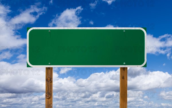 Blank green road sign with wooden posts over blue sky and clouds