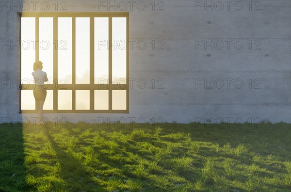Woman standing at window with bright sun light shining through concrete wall in grass and rocks field