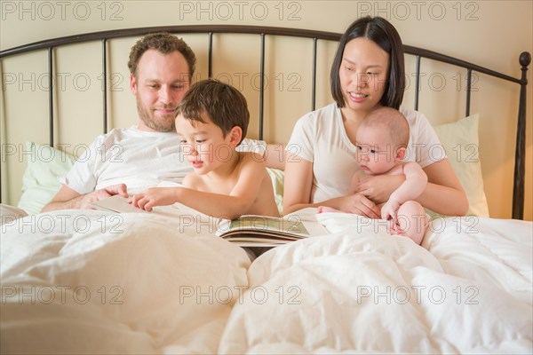 Young mixed-race chinese and caucasian baby boys reading a book in bed with their father and mother