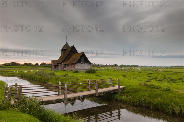 View of sheep on grazing marsh with footbridge across flooded ditch and church in evening