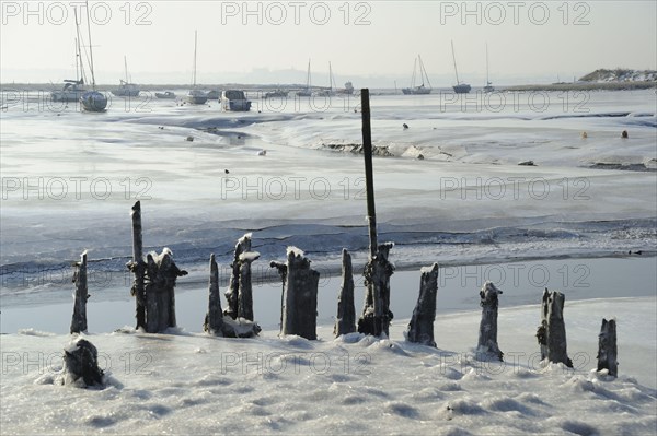 View of ice-covered mudflats and salt marshes with old jetty and sailing boats in the distance