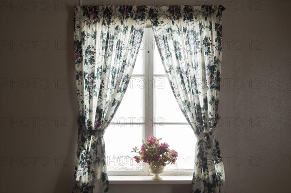 Fenster von innen mit gebluemten Gardinen und Blumenstrauss in einem alten verlassenen Haus. Vaermland