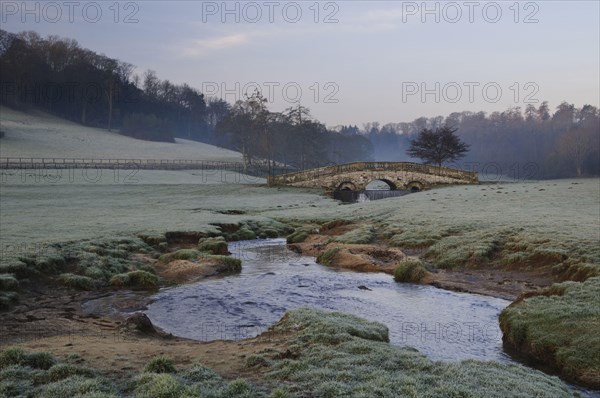 View of the arched bridge over a stream flowing through parkland at dawn