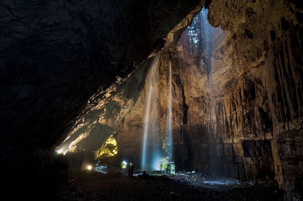 View of upland stream waterfall flowing down chasm into chamber of cave with visitors