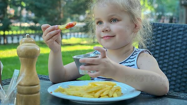 Little girl eat french fries. Close-up of blonde girl takes potato chips with her hands and tries them sitting in street cafe on the park. Odessa