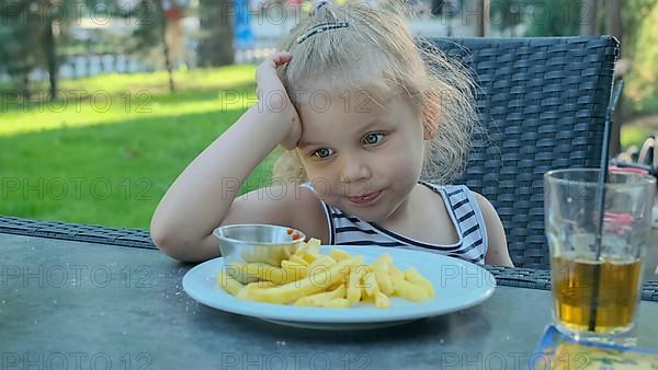 Little girl eat french fries. Close-up of blonde girl takes potato chips with her hands and tries them sitting in street cafe on the park. Odessa