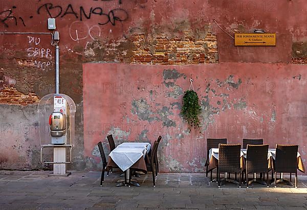 Public telephone booth and outdoor restaurant seating in front of pink house facade in the lagoon city of Venice