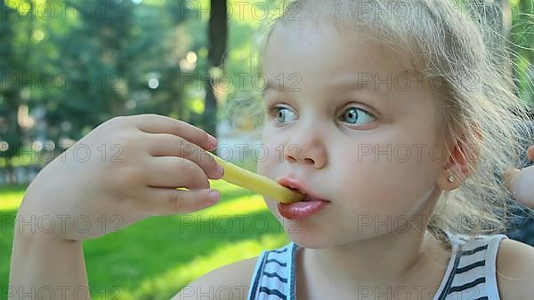 Little girl eat french fries. Close-up of blonde girl takes potato chips with her hands and tries them sitting in street cafe on the park. Odessa Ukraine