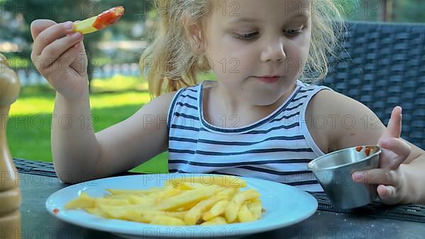 Little girl eat french fries. Close-up of blonde girl takes potato chips with her hands and tries them sitting in street cafe on the park. Odessa Ukraine