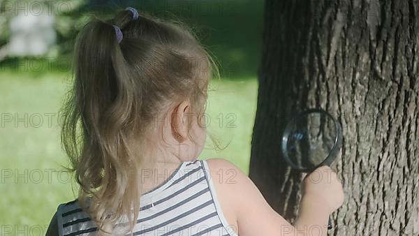 Little girl looks through the lens at insects on tree trunk. Close-up of blonde girl is studying ants while looking at them through magnifying glass on the park. Odessa Ukraine