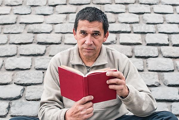 An adult man reading a red book outdoors