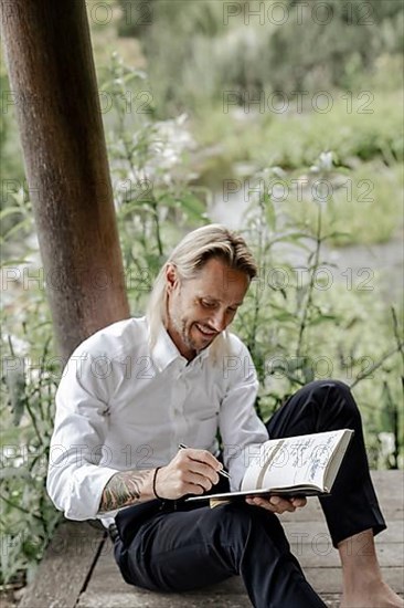 Businessman in white shirt sits on wooden jetty and writes in a book,