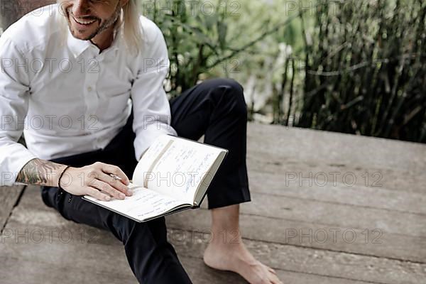 Businessman in white shirt sits on wooden jetty and writes in a book,