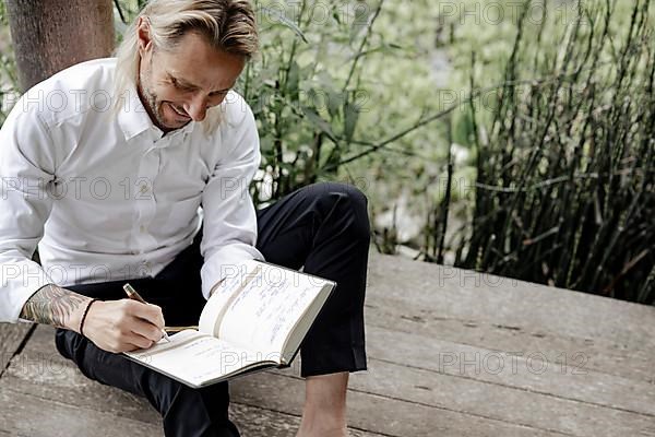 Businessman in white shirt sits on wooden jetty and writes in a book,