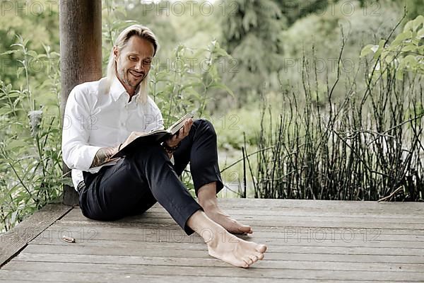 Businessman in white shirt sits on wooden jetty and writes in a book,