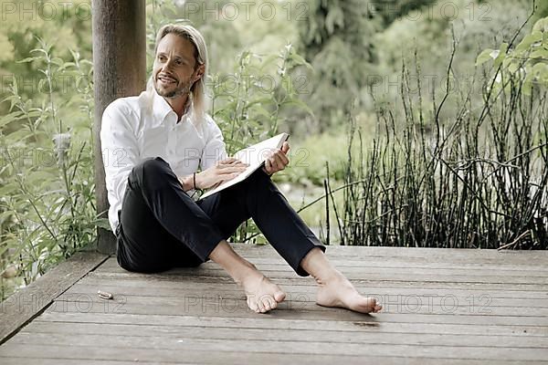 Businessman in white shirt sits on wooden jetty and writes in a book,