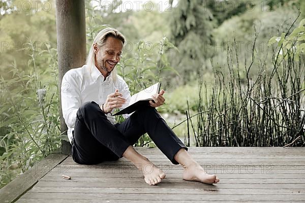 Businessman in white shirt sits on wooden jetty and writes in a book,