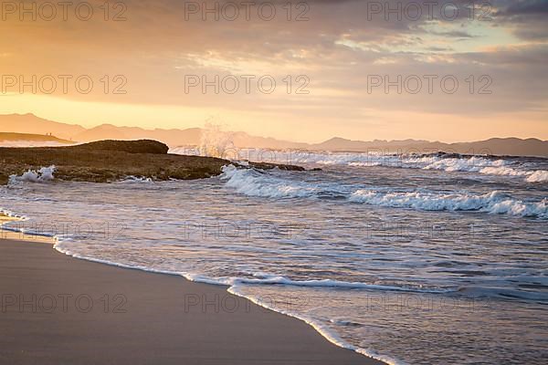 Sunset and breaking waves at the sandy beach playa in Son Serra de Marina, Tramuntana Mountains in the back