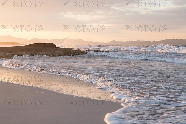 Sunset and breaking waves at the sandy beach playa in Son Serra de Marina, Tramuntana Mountains in the back