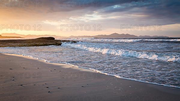 Sunset and breaking waves at the sandy beach playa in Son Serra de Marina, Tramuntana Mountains in the back