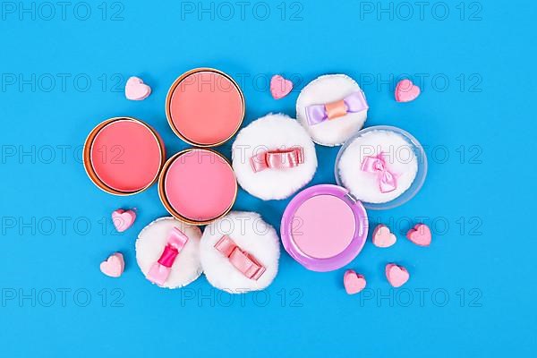 Different pink blush beauty products and powder puffs with ribbons and heart shaped pressed powder on blue background,