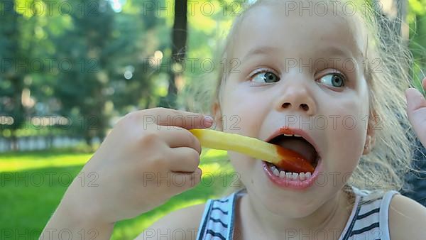 Little girl eat french fries. Close-up of blonde girl takes potato chips with her hands and tries them sitting in street cafe on the park. Odessa, Ukraine