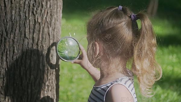 Little girl looks through the lens at insects on tree trunk. Close-up of blonde girl is studying ants while looking at them through magnifying glass on the park. Odessa, Ukraine