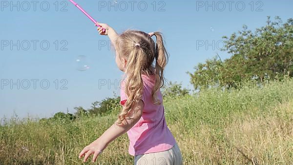 Little girl catches soap bubbles in butterfly net while playing in tall grass in park. Cute little girl smiles and catches on soap bubbles in aerial insect net in meadow on sun day. Odessa, Ukraine