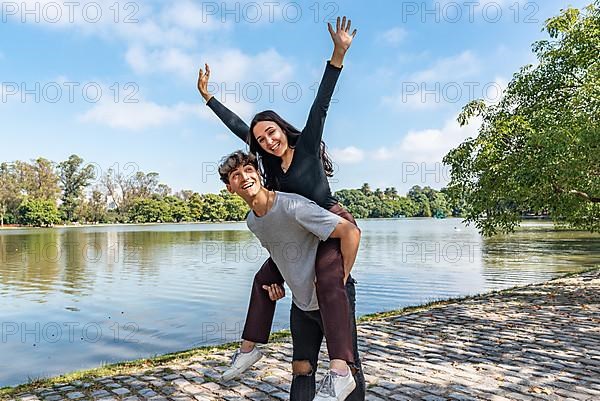 Happy couple. Handsome young man carrying his beautiful girlfriend on his back. She is raising her hands and looking at camera,