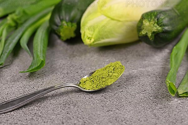 Green powder on spoon in front of raw green vegetables. Concept for natural food coloring or supplements made from vegetables,
