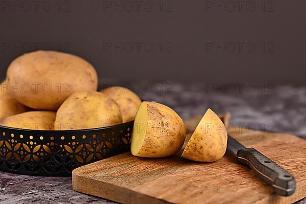 Cutting raw potato with peel on wooden cutting board with pile of whole potatoes in background,