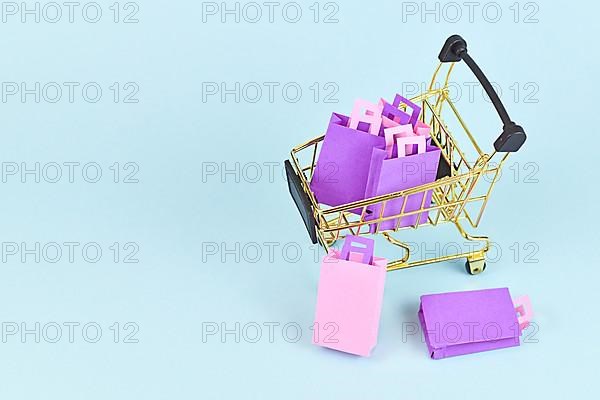 Small golden shopping cart filled with pink and purple paper shopping bags on side of blue background with copy space,