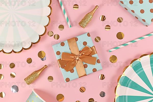 Birthday gift box with with teal blue paper plates, small golden champagne bottles and confetti on pink background