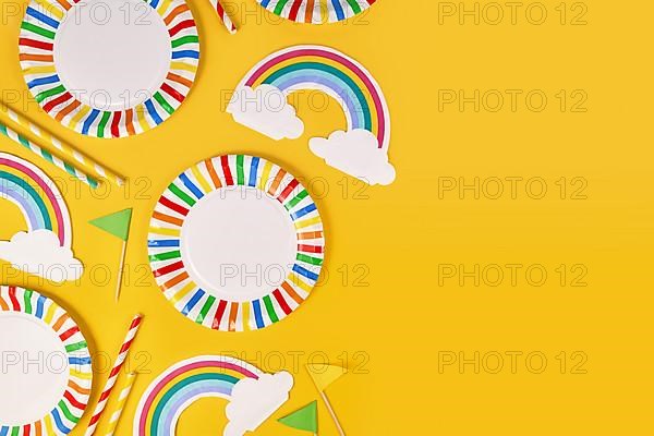 Party flat lay with colorful plates, rainbow napkins and drinking straws on yellow background with copy space