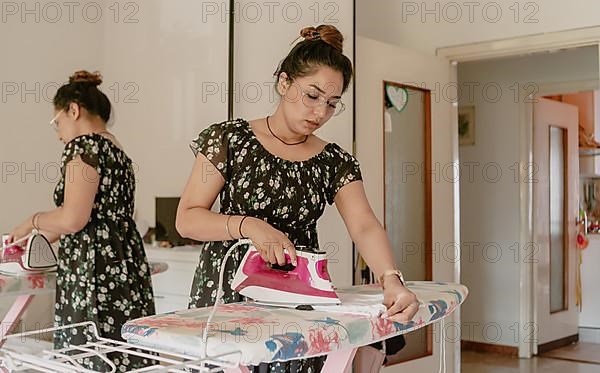 Youngwoman wearing glasses and summer silk dress with steam iron, ironing board in bed room at home in sunny day