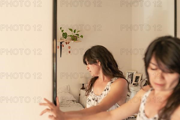 Hispanic young adult woman choosing a dress from the wardrobe in the bedroom at home, reflecting on a mirror