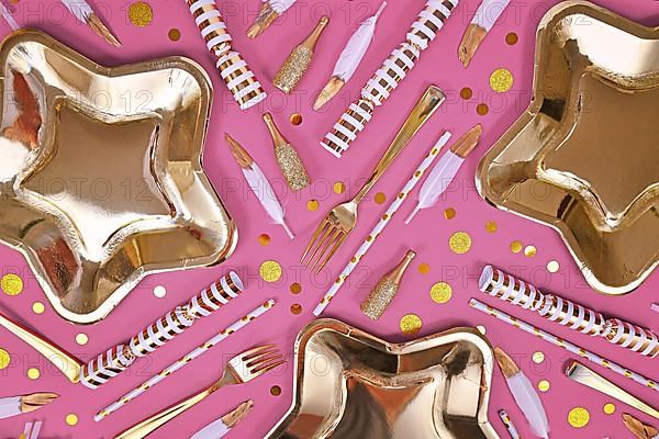 Party flat lay with golden and white decoration like plates, confetti and feathers on pink background
