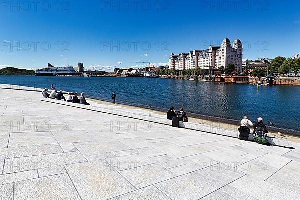 Pedestrians sitting on the white forecourt of the opera house, view from above of Oslofjord and harbour with harbour warehouse