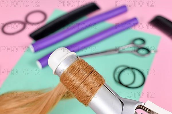 Strand of blond hair wrapped around curling iron with hair styling tools in blurry background,