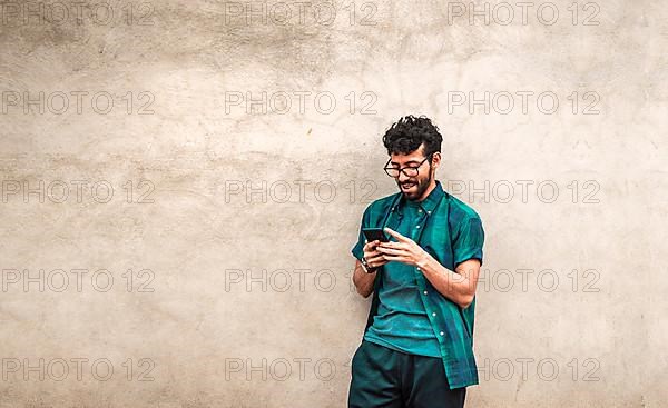 Happy handsome guy leaning against the wall using his cell phone, happy latin man with his cell phone leaning against a wall with copy space