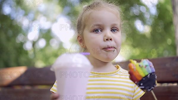 Ittle girl eats a colorful gingerbread and holds a milkshake in her hand. Close-up of cute child girl sitting on park bench and eating cookies with a milkshake and looks into the camera lens. Odessa, Ukraine