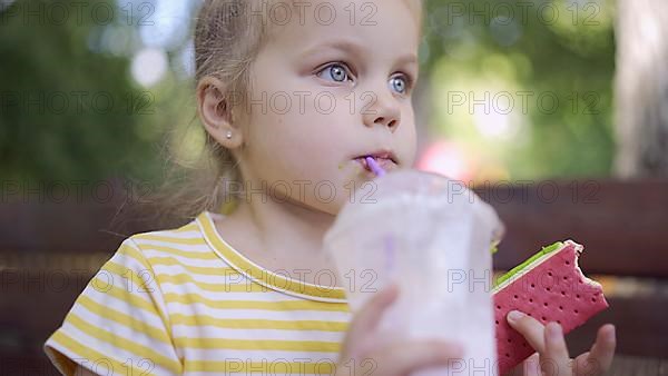 Little girl drinks a milkshake through a straw and smirks at ice cream in her other hand. Close-up portrait of cute child girl sitting on park bench and drinking milkshake. Odessa, Ukraine