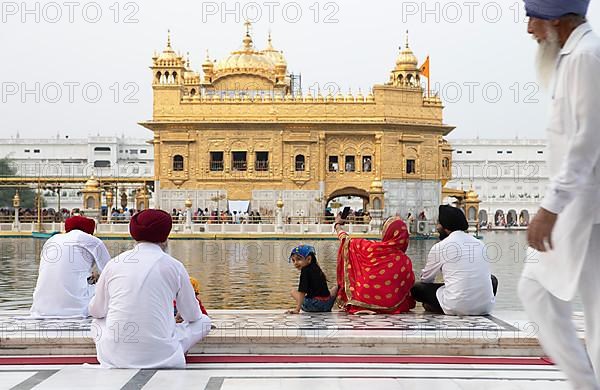 Indian woman, girl and men in white suits sitting at the edge of the water basin of the Amrit Sagar or Sacred Lake