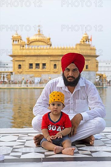 Indian Sikh in white suit and barefoot with his son sitting at the edge of the water basin of the Amrit Sagar or Sacred Lake, at the Hari Mandir or Golden Temple