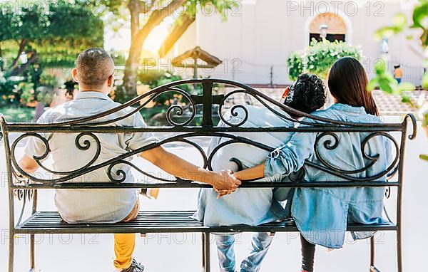 Unfaithful woman sitting holding hands with another man while boyfriend hugs her. Unfaithful couple holding hands on a park bench. Concept of love triangle and infidelity